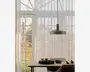 En moderne pendellampe med en bred, flad, mat metal-skærm hænger ned fra loftet i en tynd sort ledning. Lampen har et minimalistisk design og udsender et varmt, diffust lys nedad. Den er placeret over et spisebord i træ, hvorpå der står et sort serveringsfad, flere keramikkopper, en glastepotte, en stak tallerkener og en lysestage. I baggrunden ses store vinduer med hvide, lette gardiner, der lader dagslyset blødt oplyse rummet.