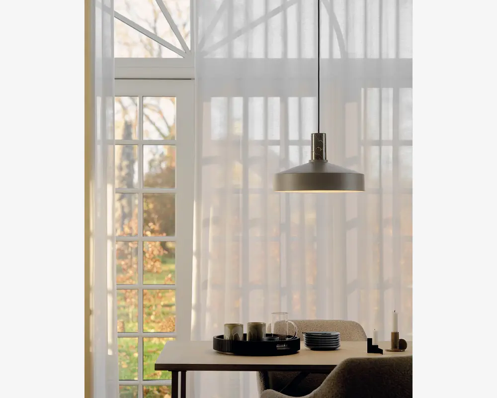 En moderne pendellampe med en bred, flad, mat metal-skærm hænger ned fra loftet i en tynd sort ledning. Lampen har et minimalistisk design og udsender et varmt, diffust lys nedad. Den er placeret over et spisebord i træ, hvorpå der står et sort serveringsfad, flere keramikkopper, en glastepotte, en stak tallerkener og en lysestage. I baggrunden ses store vinduer med hvide, lette gardiner, der lader dagslyset blødt oplyse rummet.