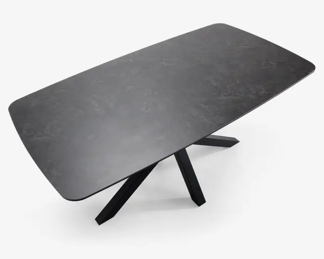 Rektangulært bord med en mørk, mat overflade, der ligner sten og har diskrete marmoreringer. Bordpladen har blødt afrundede hjørner og kanter. Understellet består af fire skråtstillede, krydsende ben i samme mørke farve, hvilket giver et moderne og solidt udtryk.