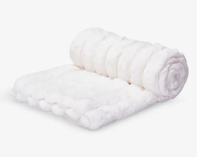 Et blødt, hvidt tæppe med en plysset og luftig tekstur. Tæppet er delvist rullet sammen, hvilket fremhæver dets tykke og hyggelige materiale samt den glatte overflade. Kanterne er pænt afsluttede, og tæppet udstråler varme og komfort – perfekt til at putte sig med eller give sengen eller sofaen et luksuriøst præg.