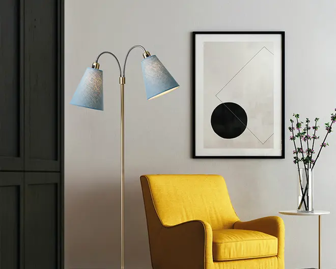 Et moderne opholdsrum med en sennepsgul lænestol, der har en blød sidde- og ryghynde, let buede armlæn og et slankt, minimalistisk design. Ved siden af stolen står en slank guldfarvet gulvlampe med to opadbuede arme, hver med en blå, kegleformet lampeskærm. Lampen står på en rund fod. Bag stolen hænger et indrammet abstrakt kunstværk med geometriske former i sort og tynde linjer på en lysegrå væg. Et lille hvidt sidebord har en klar glasvase med lange, tynde grene og små blade, som tilføjer et grønt element til rummet.