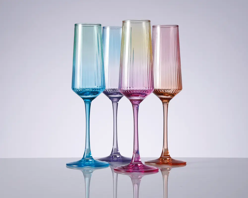 Sæt med fire høje, riflede glas i forskellige gennemsigtige farver: blå, lyse lilla, pink og rav. Hvert glas har lodrette rillede detaljer på skålen, slanke stilke og en blank overflade. Glassene står oprejst på en reflekterende overflade, hvilket fremhæver deres elegante og moderne design.