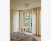 Gennemsigtige, gulvlange gardiner i en blød cremefarve hænger fra en skjult skinne og spreder blidt dagslyset i rummet. Gardinerne har et diskret lodret plisseret design, som giver et elegant og let udtryk. De er kombineret med store glasdøre og vinduer, hvilket skaber en lys og åben stemning. Stoffet virker let og halvgennemsigtigt, hvilket giver privatliv, men stadig lader dagslyset slippe ind.