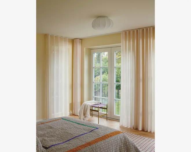 Gennemsigtige, gulvlange gardiner i en blød cremefarve hænger fra en skjult skinne og spreder blidt dagslyset i rummet. Gardinerne har et diskret lodret plisseret design, som giver et elegant og let udtryk. De er kombineret med store glasdøre og vinduer, hvilket skaber en lys og åben stemning. Stoffet virker let og halvgennemsigtigt, hvilket giver privatliv, men stadig lader dagslyset slippe ind.