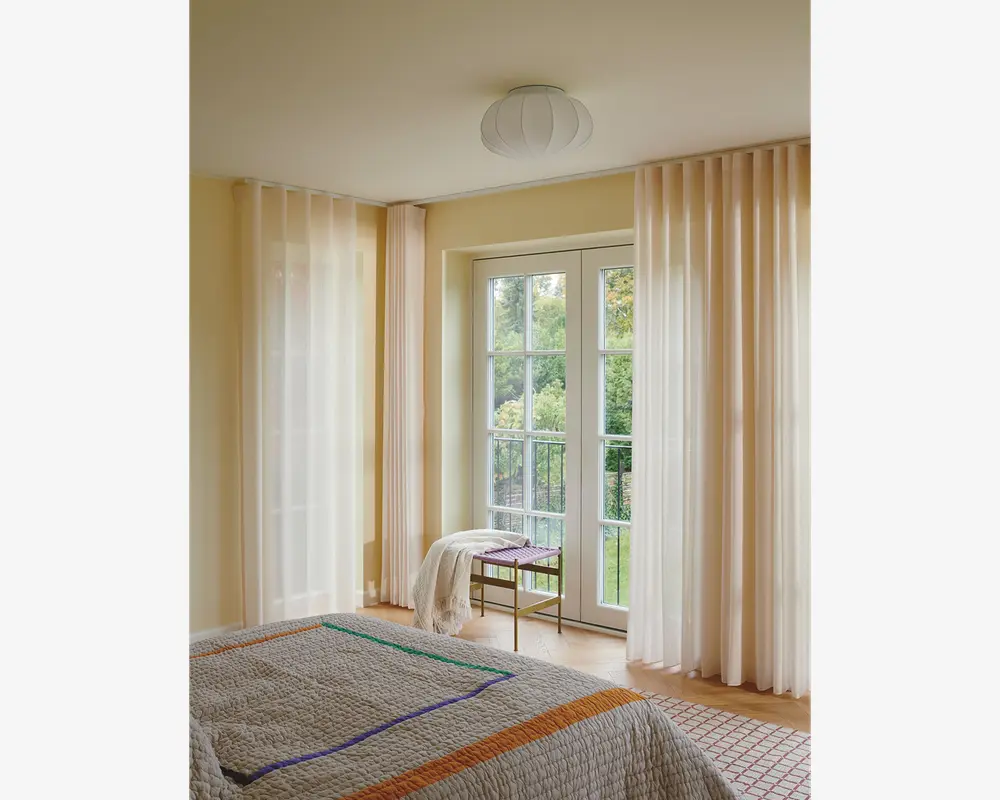 Gennemsigtige, gulvlange gardiner i en blød cremefarve hænger fra en skjult skinne og spreder blidt dagslyset i rummet. Gardinerne har et diskret lodret plisseret design, som giver et elegant og let udtryk. De er kombineret med store glasdøre og vinduer, hvilket skaber en lys og åben stemning. Stoffet virker let og halvgennemsigtigt, hvilket giver privatliv, men stadig lader dagslyset slippe ind.