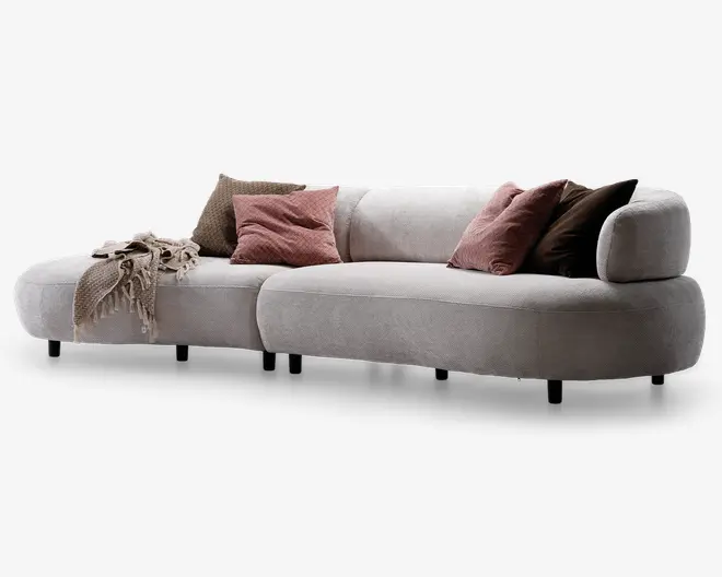 Buet, moderne sofa betrukket med lysebeige stof med lav ryg og afrundede kanter. Sofaen har en chaiselong-del til venstre. Den er pyntet med fire bløde pyntepuder i støvede rosa og mørkebrune nuancer samt et tekstureret beige tæppe, der er lagt over armlænet. Det samlede udtryk er moderne og indbydende med bløde, komfortable hynder.