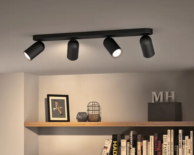 Moderne mat sort loftlampe med en lang rektangulær base og fire justerbare cylindriske spotlights, vist vinklet i forskellige retninger med varmt hvidt lys.
