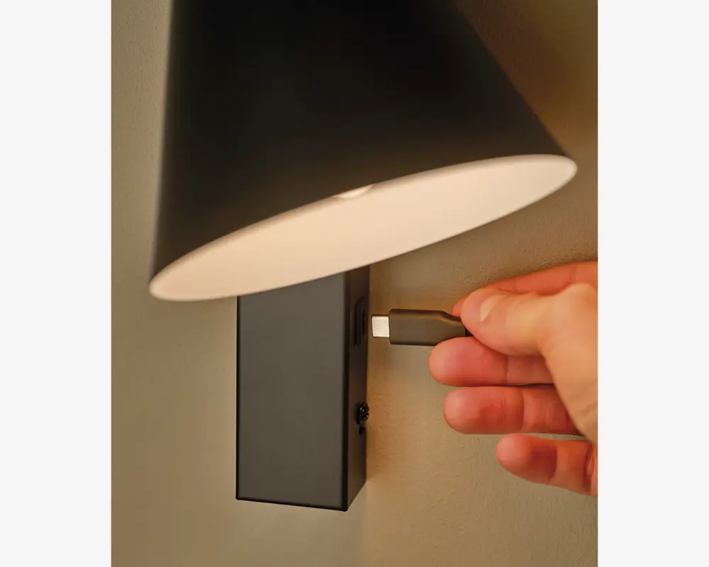 Nærbillede af en vægmonteret lampe med sort rektangulær base og konisk sort lampeskærm. Lampen har en indbygget USB-C opladningsport, hvor en hånd er ved at tilslutte et USB-C kabel. Lampen er monteret på en lys væg.