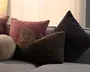 Assorterede pyntepuder i forskellige teksturer og former, herunder en støvet rosa firkantet pude, en brun krøllet rektangulær pude, en sort meleret trekantet pude og en lille beige rund pude på en grå sofa.