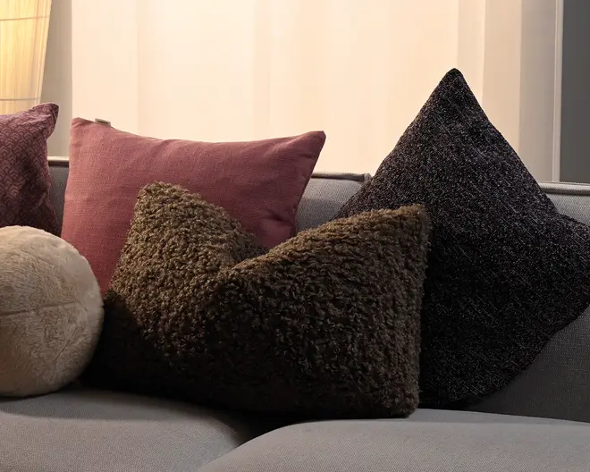 Assorterede pyntepuder i forskellige teksturer og former, herunder en støvet rosa firkantet pude, en brun krøllet rektangulær pude, en sort meleret trekantet pude og en lille beige rund pude på en grå sofa.