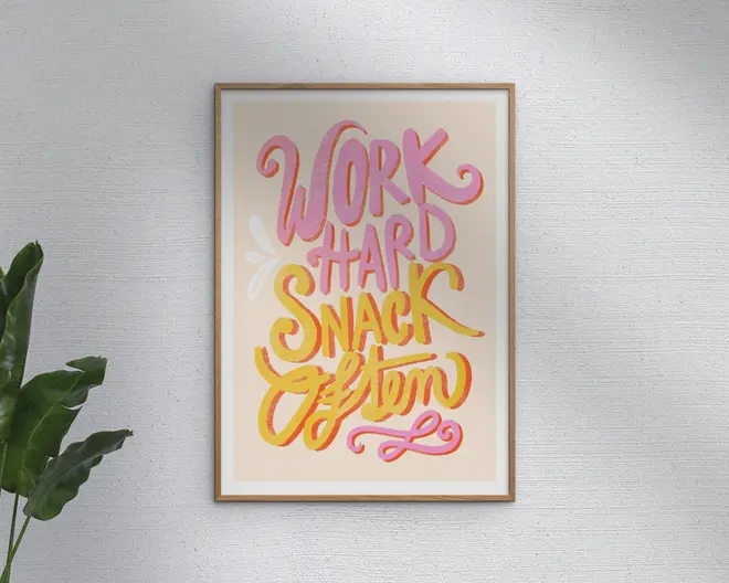 Et indrammet print med den motiverende sætning 'Work Hard Snack Often' skrevet med store, legende bogstaver. Teksten er i nuancer af pink, gul og orange på en lys beige baggrund, med en diskret dekorativ detalje. Rammen er enkel og i lyst træ, og printet hænger på en hvid, tekstureret væg med en grøn plante delvist synlig til venstre.