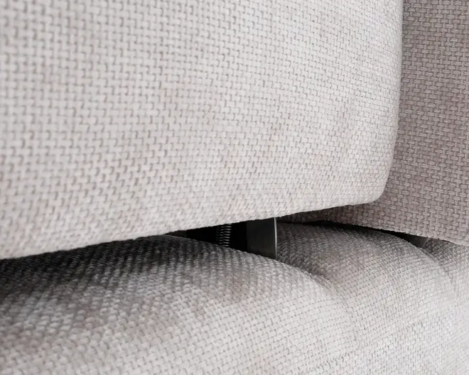 Nærbillede af en lys beige, tekstureret stofpolstring med et synligt mellemrum mellem to polstrede sektioner. Stoffet har et vævet mønster, og der ses et metalhængsel eller støttebeslag i mellemrummet, hvilket indikerer justerbar eller tilbagelænet funktion.