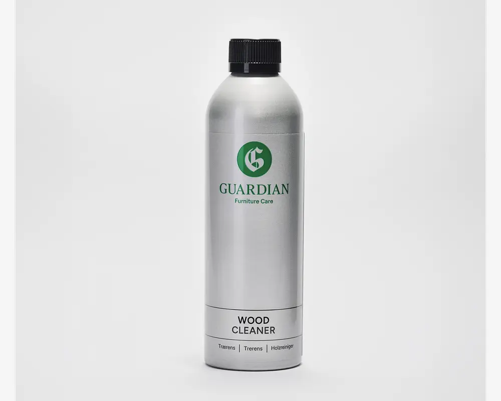 En høj, cylindrisk sølvfarvet flaske med sort skruelåg. Etiket med grønt 'G'-logo og mærket 'Guardian Furniture Care'. Produktet er angivet som trærenser, med yderligere tekst på flere sprog nederst. Flasken har et slankt, moderne design og ser ud til at være lavet af metal.