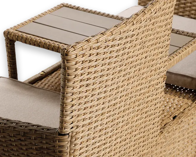 Nærbillede af et udendørsmøbel med tæt flettet syntetisk rattan i en naturlig beige farve. Armlænet har et integreret sidebord med en lys, lamelagt overflade. Sædet har en tyk, lys beige hynde for ekstra komfort. Konstruktionen virker solid og vejrbestandig, velegnet til terrasse eller have.