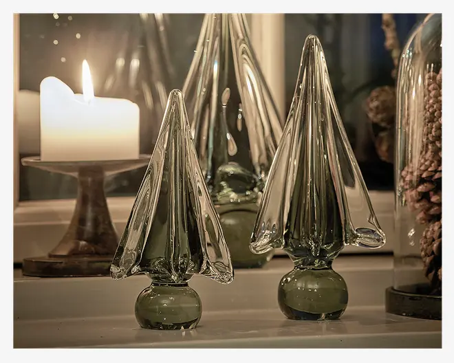 Sæt af dekorative glasskulpturer formet som stiliserede træer, lavet af klart glas med et diskret grønt skær. Hver figur har en glat, rund fod og lange, spidse grene, hvilket giver et moderne og elegant udtryk. Glasset er blankpoleret og reflekterer lyset flot. Produkterne er udstillet på en overflade med et tændt stearinlys i baggrunden, hvilket skaber en varm og hyggelig stemning.