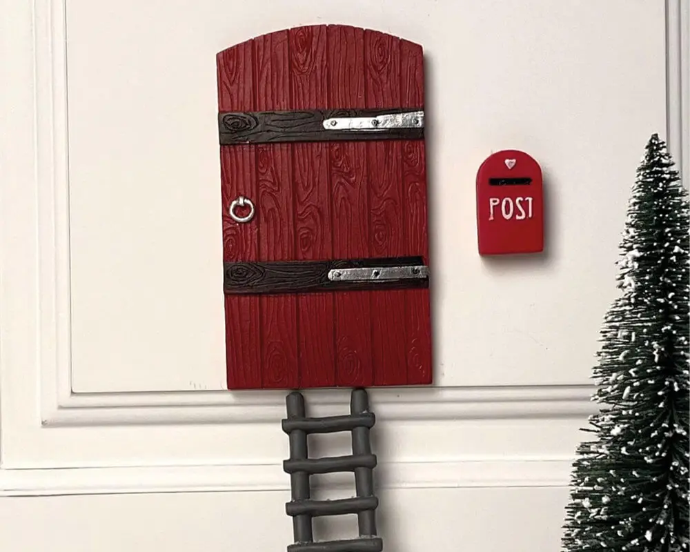 En lille, dekorativ rød dør med træstruktur og to vandrette mørkebrune bjælker, hver med en sølvfarvet metalbeslag. Døren har et rundt metalringhåndtag i venstre side. Ved siden af døren hænger en miniature rød postkasse med teksten 'POST' i hvide bogstaver. Under døren er der en kort, grå stige. Til højre står et lille kunstigt grantræ med hvidt på spidserne, som ligner sne.