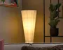 Høj, konisk gulvlampe med en cremefarvet stofskærm, der lyser varmt og skaber et blødt stemningslys. Vist i en hyggelig stue ved siden af en potteplante og bag en sofa.