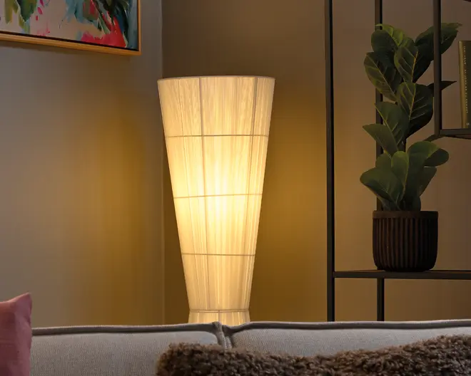 Høj, konisk gulvlampe med en cremefarvet stofskærm, der lyser varmt og skaber et blødt stemningslys. Vist i en hyggelig stue ved siden af en potteplante og bag en sofa.
