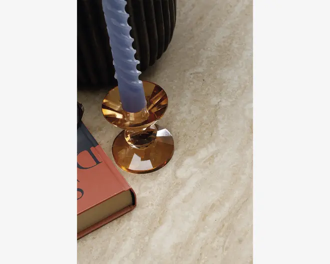 Billedet viser en nærbillede af en beige, marmoreret overflade. På overfladen står en facetslebet lysestage i ravfarvet glas med et lyse lilla snoet stagelys. Ved siden af lysestagen ligger en hardback-bog med rød og blå bogomslag, og en del af en mørk, rillet keramikvase kan ses i baggrunden.