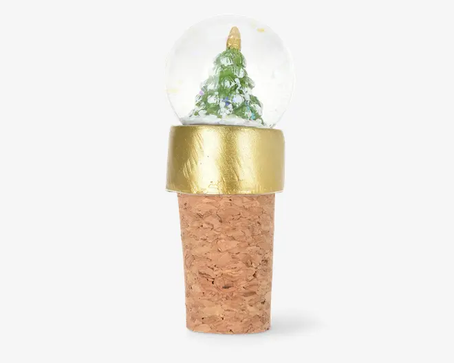 Flaskestopper af kork med en dekorativ glaskugle på toppen, som indeholder et miniature juletræ med sne-lignende detaljer. Glaskuglen er indfattet i et guldfarvet metalbånd, hvilket giver et festligt og elegant udtryk. Korkbasen er naturlig og struktureret, designet til at passe sikkert i flaskehalsen.