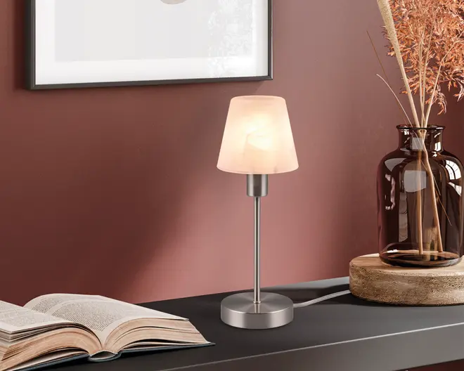 En lille bordlampe med en fod i børstet metal og en slank, lige stang. Lampen har en matteret glasskærm med et diskret marmoreret mønster, der giver et blødt og diffust lys. Designet er minimalistisk og moderne, velegnet til skriveborde eller sengeborde. Lampen er tilsluttet strøm, og ledningen er synlig fra lampens fod.