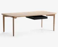 Rektangulært bord med lys træfinish, afrundede hjørner og slanke, let tilspidsede ben. Under bordpladen er der en enkelt sort udtræksskuffe placeret på den ene side, som giver diskret opbevaringsplads. Designet er minimalistisk og moderne med glatte overflader og rene linjer.