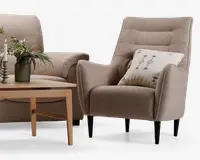 En moderne lænestol betrukket med lysebrunt stof og en let buet ryg med vandrette syninger, der giver ekstra struktur. Stolen har slanke, koniske sorte ben og leveres med en dekorativ pude med broderet geometrisk mønster i afdæmpede farver. Armlænene er let udadbuede, hvilket giver en behagelig siddeoplevelse.