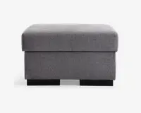 Rektangulær puf med blød, polstret top og betrukket med et tekstureret lysegråt stof. Designet er enkelt og moderne med rene linjer og en firkantet form. Puffen står på lave, solide sorte ben, og stoffet er stramt monteret for et pænt udseende.
