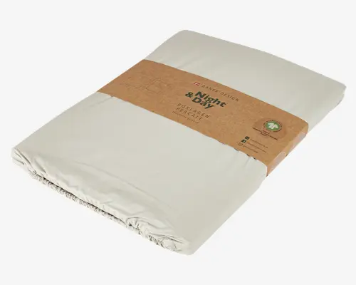 Lys beige faconsyet lagen i glat percale-bomuld, pænt sammenfoldet og omviklet med et brunt bånd af genbrugspapir. Emballagen fremhæver dansk design, miljøvenligt materiale og elastikkant, der sikrer en tæt pasform omkring madrassen.