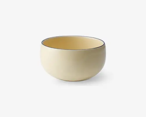 Rund skål med en glat, mat cremefarvet yderside og en matchende inderside. Skålens kant er fremhævet med en tynd, mørk streg, hvilket giver et moderne og minimalistisk udtryk. Skålen har et enkelt, ensartet design uden hank eller mønstre.