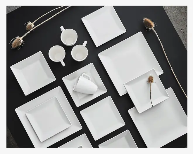 Et sæt moderne, minimalistisk hvidt stel med firkantede og rektangulære tallerkener i forskellige størrelser samt matchende hvide kopper og underkopper. Tallerkenerne har rene, skarpe kanter og en glat, blank overflade. Kopperne har et enkelt håndtag og et stilrent design. Opstillingen fremhæver sættets ensartethed og elegance, velegnet til et moderne spisebord.