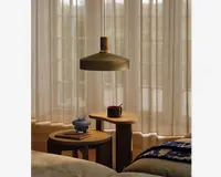 To runde sideborde i træ med forskellige højder står ved siden af hinanden. På det højere bord står et mørkt keramikkrus med en ske, mens det lavere bord har en lille blå vase og en lukket notesbog med en pen ovenpå. Bordene har et minimalistisk design med tykke, solide ben. En stor pendellampe med mat finish hænger over bordene og giver et varmt lys. I baggrunden dækker lyse, gulvlange gardiner store vinduer, så blødt dagslys filtreres ind i rummet. Foran ses en del af en seng med et blåt og hvidt mønstret tæppe.