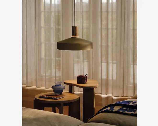 To runde sideborde i træ med forskellige højder står ved siden af hinanden. På det højere bord står et mørkt keramikkrus med en ske, mens det lavere bord har en lille blå vase og en lukket notesbog med en pen ovenpå. Bordene har et minimalistisk design med tykke, solide ben. En stor pendellampe med mat finish hænger over bordene og giver et varmt lys. I baggrunden dækker lyse, gulvlange gardiner store vinduer, så blødt dagslys filtreres ind i rummet. Foran ses en del af en seng med et blåt og hvidt mønstret tæppe.