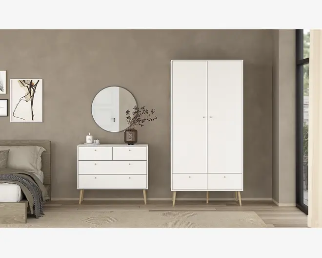 Et moderne, minimalistisk soveværelsesmiljø med en hvid kommode, der har tre store skuffer og to mindre øverste skuffer, alle med små runde greb. Kommoden står på skråtstillede ben i lyst træ. Ved siden af står et matchende hvidt garderobeskab med to låger og to nederste skuffer, også med små runde greb og ben i lyst træ. Møblerne har et rent, enkelt design med glatte overflader og diskrete detaljer. Over kommoden er der monteret et rundt spejl, og på toppen står en dekorativ vase med grene. Det samlede udtryk er stilrent og moderne med neutrale farver og naturlige træelementer.