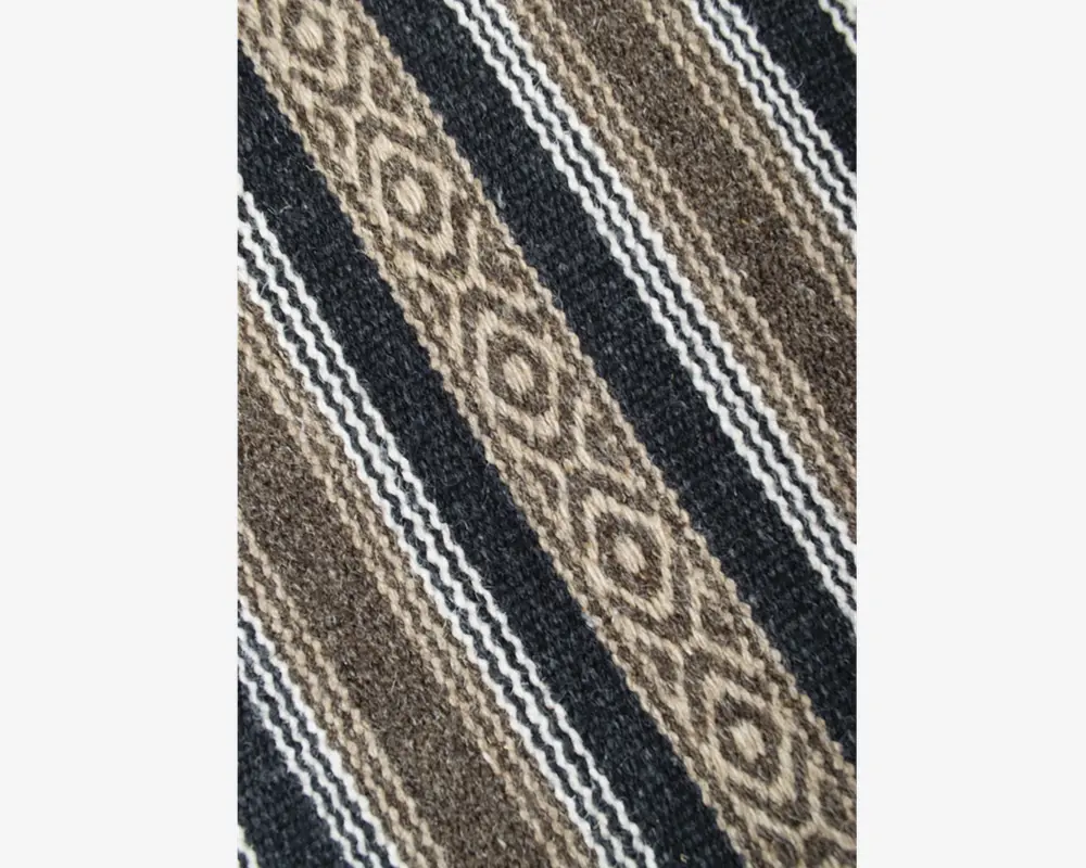 Nærbillede af et vævet tekstil med diagonale striber i brune, beige, sorte og hvide nuancer. Mønstret består af skiftevis ensfarvede og teksturerede striber, hvor nogle striber har diamantformede, geometriske motiver. Stoffet ser tykt og slidstærkt ud med en let grov overflade.