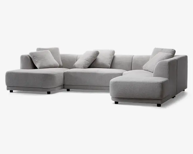 En moderne, L-formet sofa med lysegråt stofbetræk. Sofaen har brede, dybe sæder og lave ryglæn, hvilket giver et moderne udtryk. Den leveres med fire matchende, firkantede puder for ekstra komfort. Designet har rene linjer og blokformede armlæn, hvor den ene side er forlænget som en chaiselong for ekstra afslapningsplads.