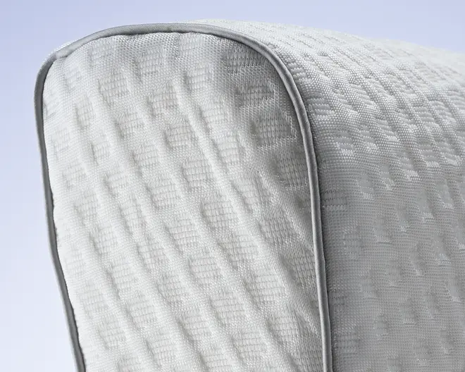 Nærbillede af et lysegråt stof med et diskret geometrisk teksturmønster. Stoffet er tæt vævet og har et hævet kvadratisk design, hvilket giver det et moderne og taktilt udtryk. Kanten er afsluttet med en glat, rund piping i samme farve, hvilket giver en pæn og holdbar syning.