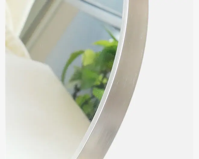 Nærbillede af et rundt spejl med en elegant ramme i børstet metal. Rammen har en glat, let mat overflade. Spejlets refleksion viser en sløret baggrund med grønne stueplanter og et vindue, hvilket antyder et moderne indendørs miljø.