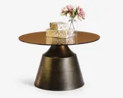 Rundt bord med en reflekterende bronzefarvet glasplade og en bred, konisk fod i børstet metal. Bordet er pyntet med en klar glasvase med lyserøde kunstige blomster og tre stablede gaveæsker med guldfarvet blomstermønster.