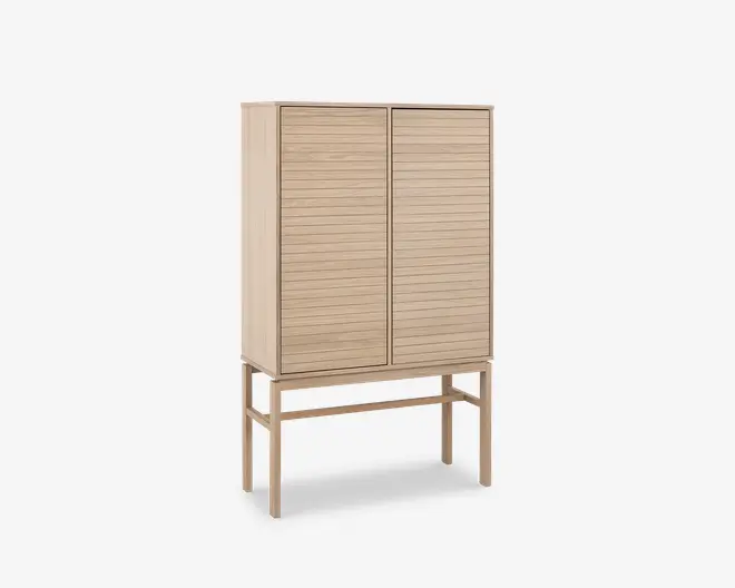 Høj skab lavet af lyst træ med to låger, der har vandrette lameldetaljer. Skabet står på fire slanke ben, som er forbundet med støttebøjler, hvilket giver et løftet og let udtryk. Designet er minimalistisk og moderne med rene linjer og en naturlig træfinish.