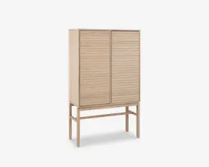 Høj skab lavet af lyst træ med to låger, der har vandrette lameldetaljer. Skabet står på fire slanke ben, som er forbundet med støttebøjler, hvilket giver et løftet og let udtryk. Designet er minimalistisk og moderne med rene linjer og en naturlig træfinish.
