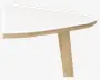 Nærbillede af et bordhjørne med en hvid, glat og afrundet bordplade med synlig lagdelt trækant. Bordet har et skråt ben i lyst træ, hvilket understreger et moderne og minimalistisk design.