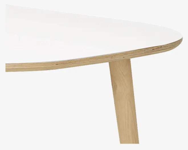 Nærbillede af et bordhjørne med en hvid, glat og afrundet bordplade med synlig lagdelt trækant. Bordet har et skråt ben i lyst træ, hvilket understreger et moderne og minimalistisk design.