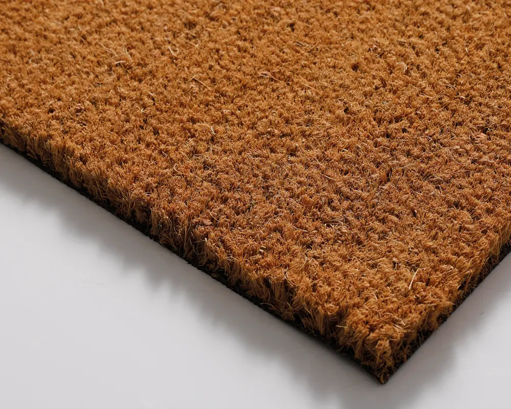 Nærbillede af en rektangulær måtte lavet af grove, naturlige brune fibre. Overfladen har en ru, tekstureret struktur, der er velegnet til at skrabe snavs af sko. Måtten er forholdsvis tyk med tætpakkede børster og en solid, flad bund for stabilitet på gulvet.