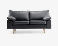 En moderne to-personers sofa betrukket med glat sort læder og udstyret med bløde, aftagelige ryg- og sædehynder. Sofaen har brede, polstrede armlæn og står på fire lyse træben, hvilket giver et nutidigt udtryk. Designet er minimalistisk, og materialerne tyder på nem vedligeholdelse og god komfort.