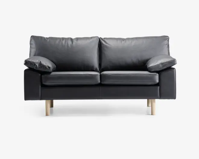 En moderne to-personers sofa betrukket med glat sort læder og udstyret med bløde, aftagelige ryg- og sædehynder. Sofaen har brede, polstrede armlæn og står på fire lyse træben, hvilket giver et nutidigt udtryk. Designet er minimalistisk, og materialerne tyder på nem vedligeholdelse og god komfort.