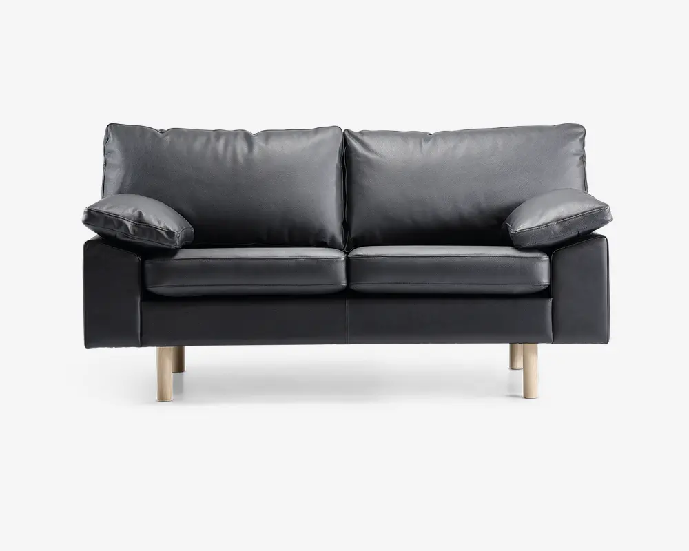 En moderne to-personers sofa betrukket med glat sort læder og udstyret med bløde, aftagelige ryg- og sædehynder. Sofaen har brede, polstrede armlæn og står på fire lyse træben, hvilket giver et nutidigt udtryk. Designet er minimalistisk, og materialerne tyder på nem vedligeholdelse og god komfort.