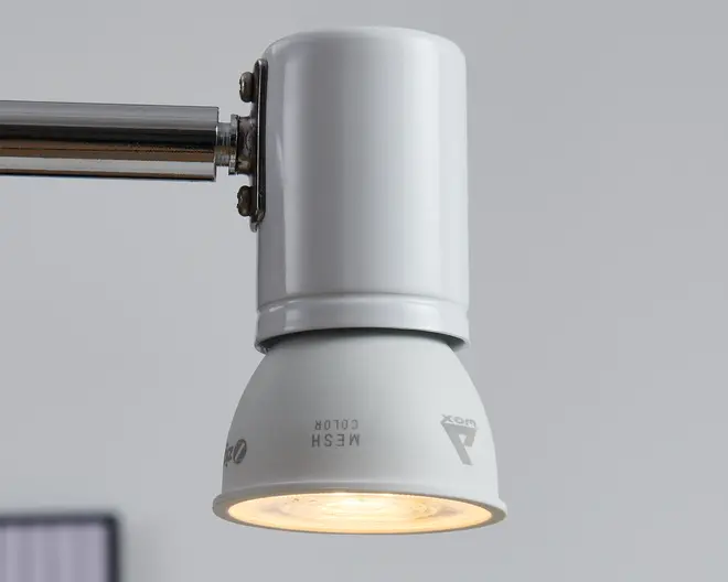 En hvid, cylindrisk og kegleformet justerbar lampe monteret på en vandret kromstang, med et varmt lys, der skinner nedad fra undersiden.