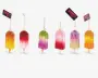 Et sæt med seks pynt formet som ispinde, hver med en farverig gradient i nuancer som gul, orange, pink, lilla og grøn. Toppen af hver pynt er dekoreret med glimmer i matchende eller kontrastfarver, hvilket giver en glitrende effekt. Hver pynt hænger i en gylden snor og har et lille mærke vedhæftet. Ispindene har en realistisk træpind i bunden, og overfladen er blank og festlig.