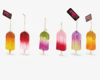 Et sæt med seks pynt formet som ispinde, hver med en farverig gradient i nuancer som gul, orange, pink, lilla og grøn. Toppen af hver pynt er dekoreret med glimmer i matchende eller kontrastfarver, hvilket giver en glitrende effekt. Hver pynt hænger i en gylden snor og har et lille mærke vedhæftet. Ispindene har en realistisk træpind i bunden, og overfladen er blank og festlig.